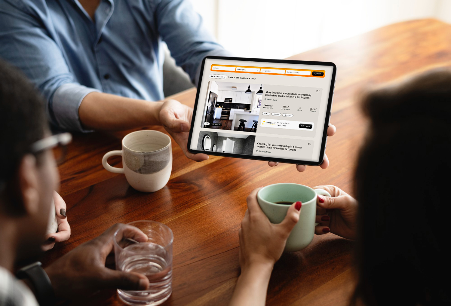 Three people sitting at a wooden table, looking at a tablet displaying real estate listings.